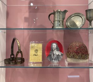 A display of historic items on a shelf against a mauve backdrop. On the left is a metal object made up of a band and a loop on the right is a red silk hat decorated with fine gold details. 