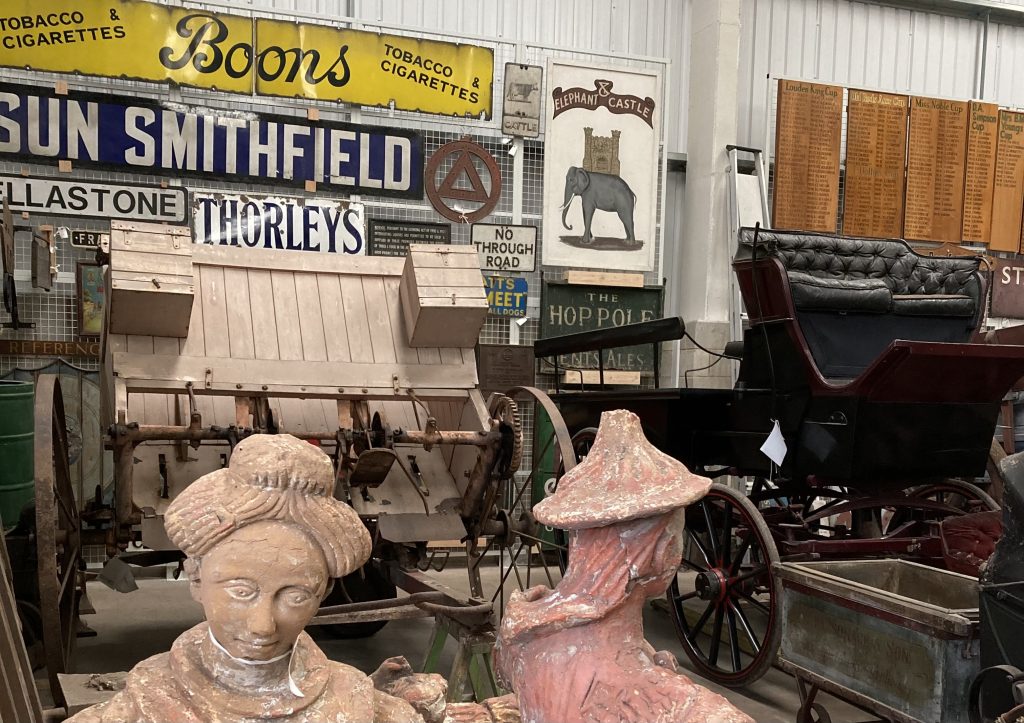 A collection of museum objects in a storeroom. There are colourful metal signs in the background, a cart and a carriage with black leather seats. In the foreground are two terracotta statues.
