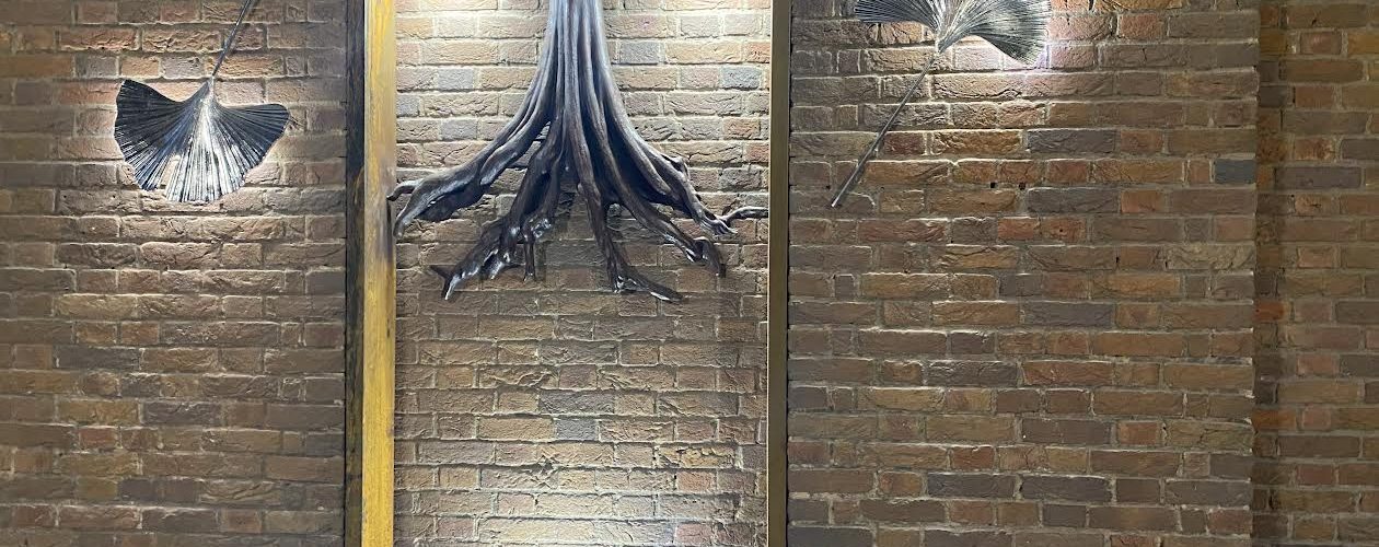 A metal light art sculpture fixed to a re dbrick wall. Leaves and tree roots are sculpted from metal and illuminated with white spotlights.