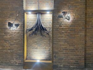 A metal light art sculpture fixed to a re dbrick wall. Leaves and tree roots are sculpted from metal and illuminated with white spotlights.